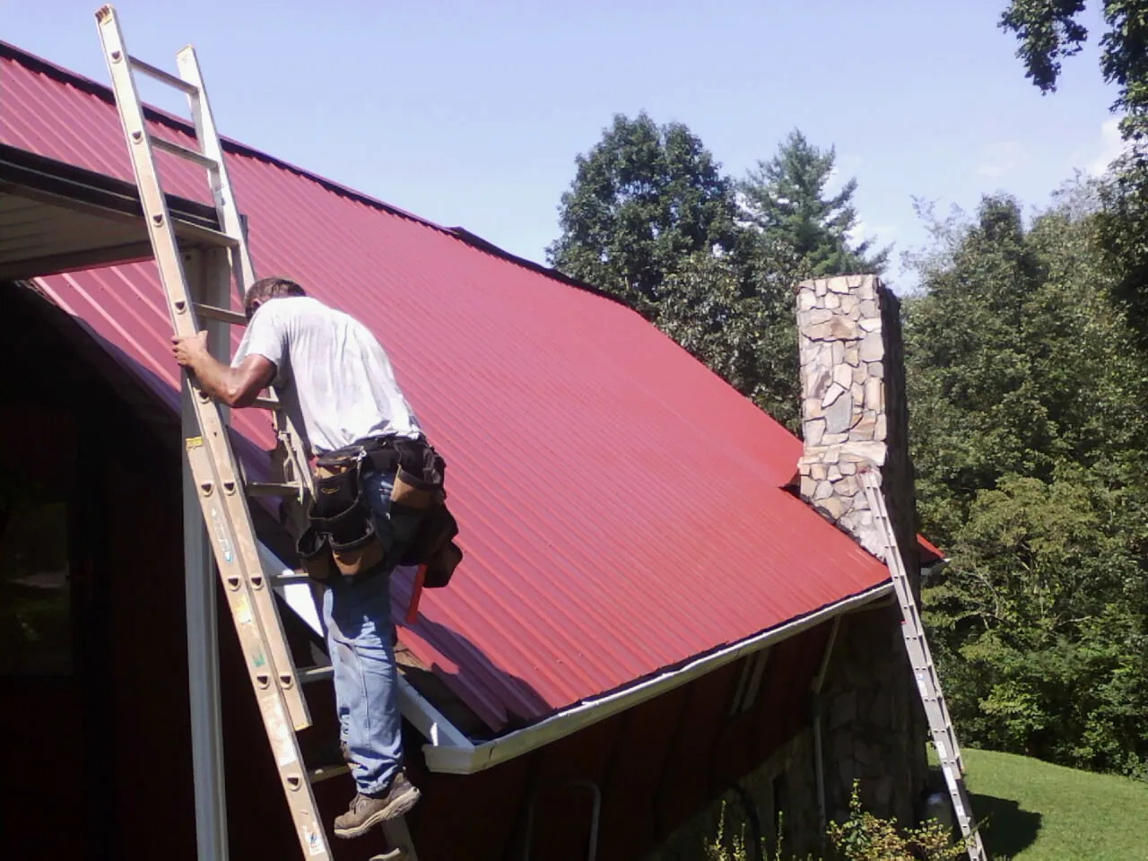 Roofer on a ladder beside a red metal roof and stone chimney — Soffit & Fascia Repair by Roofing Brothers in Yoder